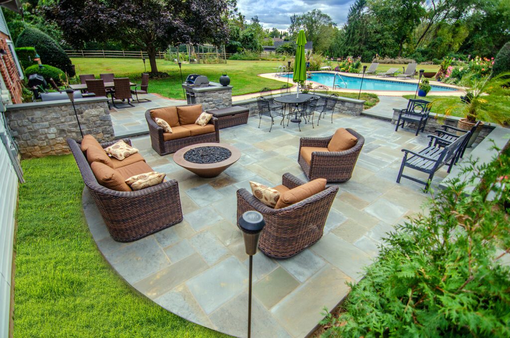 A stone patio with a round fire table and wicker furniture with cushions surrounding it.