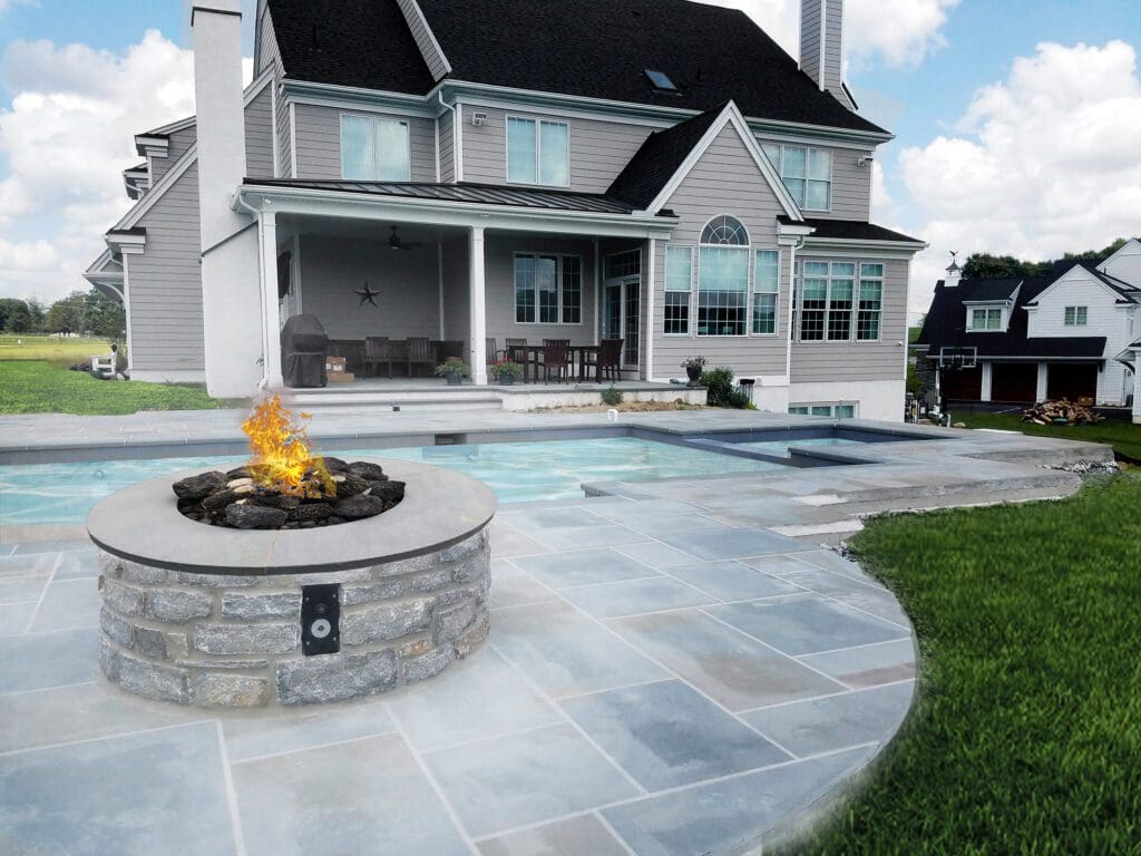 A propane fire pit burns on a flagstone patio next to a backyard pool.