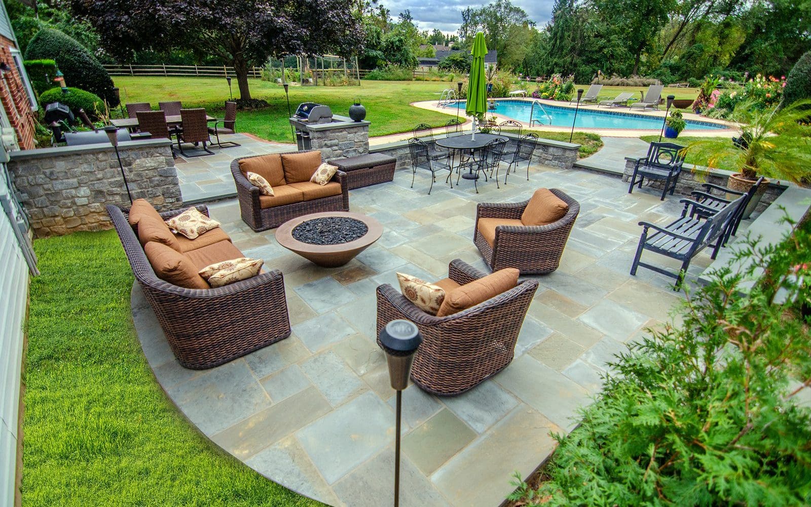 A stone patio with a round fire table and wicker furniture with cushions surrounding it.
